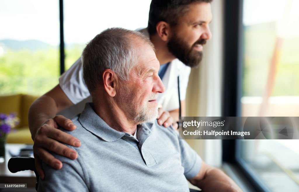 A male healthcare worker talking to senior man in wheelchair indoors at home.