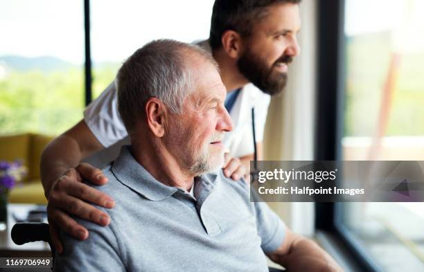 a male healthcare worker talking to senior man in wheelchair indoors at home. - doctor with patient man stockfoto's en -beelden