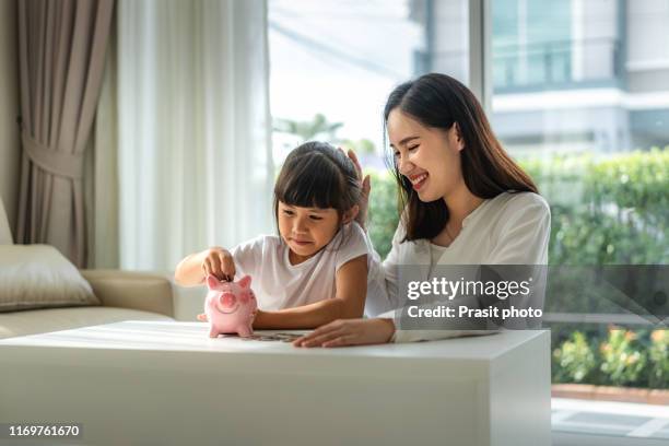 happy mother and daughter saving money putting coins into piggy bank on table at home. - family savings stock pictures, royalty-free photos & images