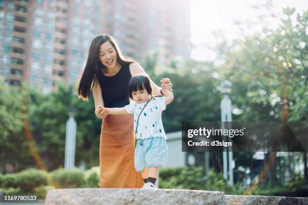 joyful young asian mother holding hands of little daughter supporting and assisting her walking along on stone on a lovely sunny day - china balance stock pictures, royalty-free photos & images