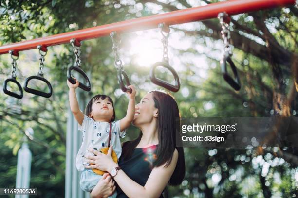 young asian mother supporting her little daughter while playing with the monkey bars at the outdoor playground on a lovely sunny day - klettern park stock-fotos und bilder