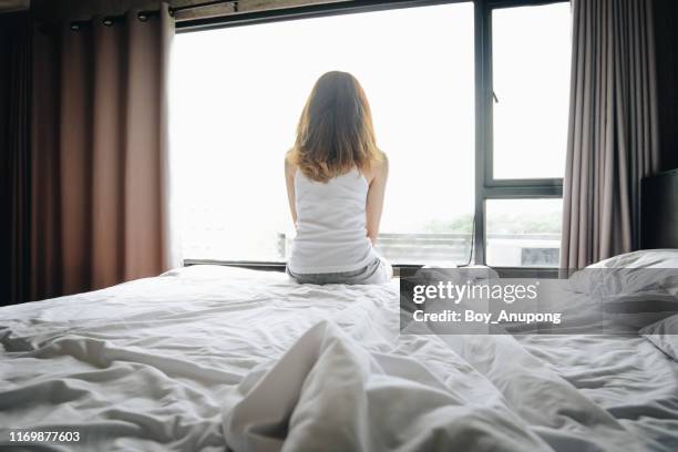 portrait of depressed woman sitting alone on bed, looking to outside the window. - indecisive woman stock pictures, royalty-free photos & images