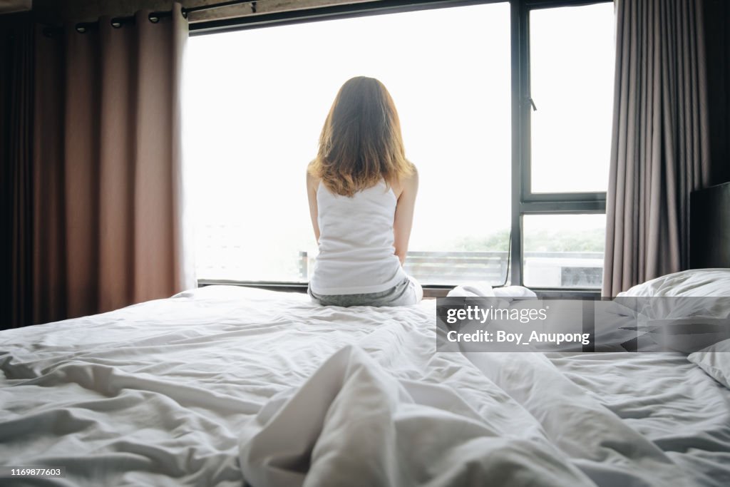 Portrait of depressed woman sitting alone on bed, looking to outside the window.