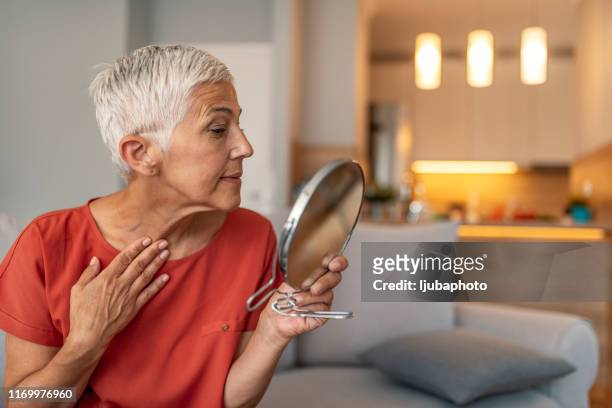 mujer madura con espejo de maquillaje masajeando su cara y cuello - wrinkles fotografías e imágenes de stock