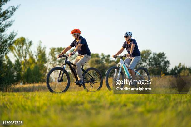 active senior couple cycling on mountain bikes through rural landscape - 2018 cycling stock pictures, royalty-free photos & images