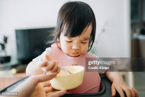 asian toddler girl sitting on high chair feeding herself at home - bib stock pictures, royalty-free photos & images