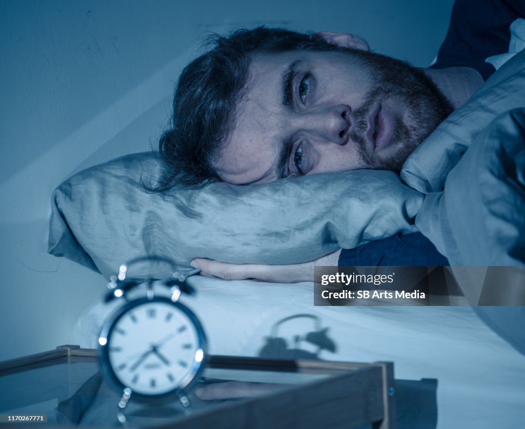 Schlafloser und verzweifelter junger kaukasischer Mann erwacht nachts nicht in der Lage zu schlafen, fühlte sich frustriert und besorgt Blick auf die Uhr leidet an Schlaflosigkeit im Stress und Schlafstörungen Konzept.