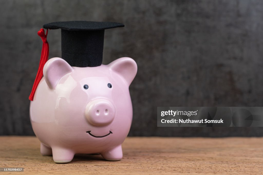 Smiling pink piggy bank wearing graduated hat on wooden table with dark black background and copy space, education fund, Scholarships, university cost and expense or saving for student loan concept