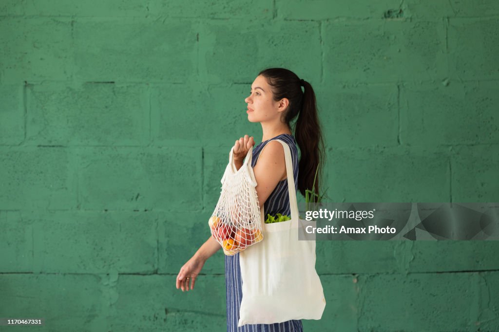 Jeune femme avec le sac d'Eco de coton d'achat avec des fruits et légumes dans ses mains sur le fond vert de mur. Mode de vie, concept zéro déchet