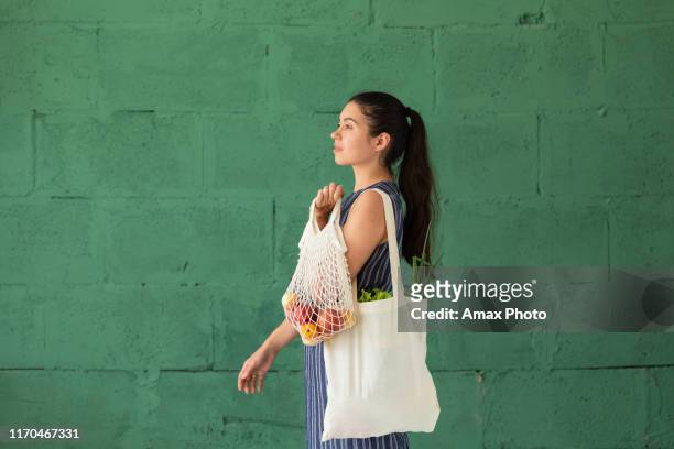 jeune femme avec le sac d'eco de coton d'achat avec des fruits et légumes dans ses mains sur le fond vert de mur. mode de vie, concept zéro déchet - sac à main blanc photos et images de collection