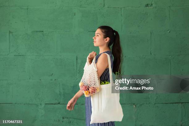 young woman with shopping cotton eco bag with fruits and vegetables in her hands on green wall background. lifestyle, zero waste concept - consumerism imagens e fotografias de stock