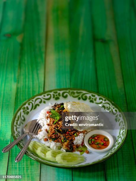 das berühmte würzige thai-gericht namens "pak krapao moo", gebratenes schweinefleisch mit heiligem basilikum auf englisch, serviert auf einem bett aus gedämpftem reis mit einem spiegelei, gurke und "pik nam bla" (chili mit fischsauce) auf einem alten gr - nuoc cham stock-fotos und bilder