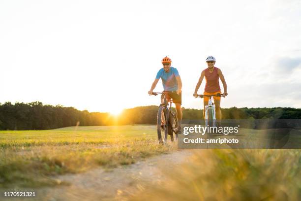 happy mature adult sporty couple on mountainbikes in rural landscape at sunset - nature field trip stock pictures, royalty-free photos & images