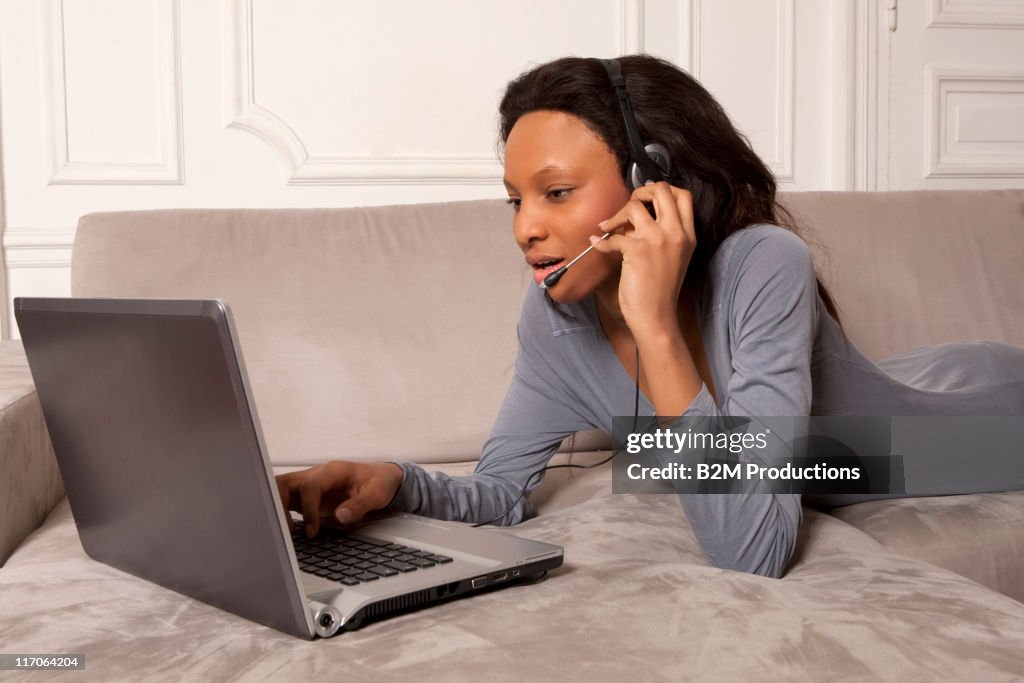 Woman with headset lying on sofa using laptop