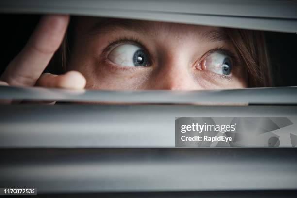 terrified wide-eyed teenage girl looking through closed blinds - fobia imagens e fotografias de stock