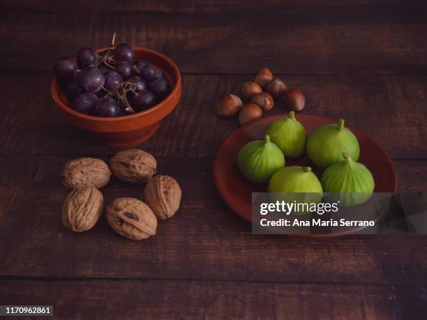 autumn fruits: white grapes in a clay bowl, freshly harvested green figs in a clay plate and hazelnuts and nuts on an old dark wooden table - fig stock pictures, royalty-free photos & images