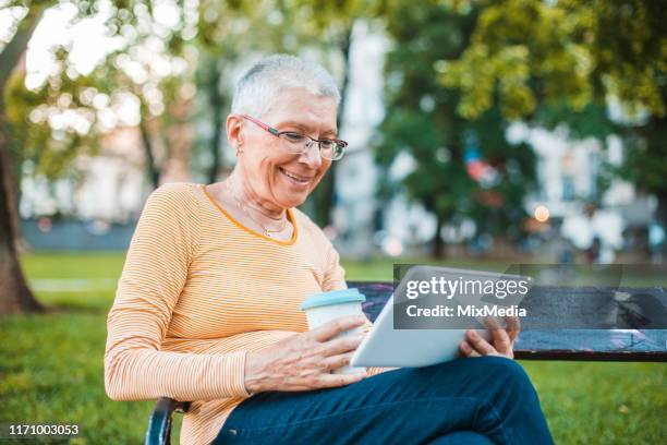 seniorin sitzt im park, trinkt kaffee und benutzt ein digitales tablet - coole oma stock-fotos und bilder