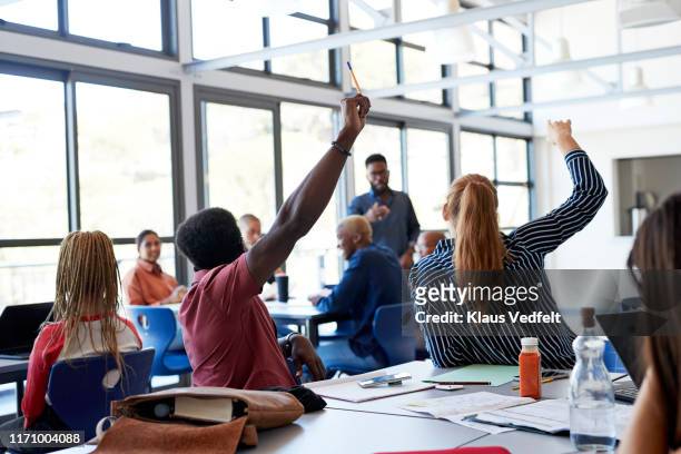 multi-ethnic students raising hands at desk - participant stock pictures, royalty-free photos & images