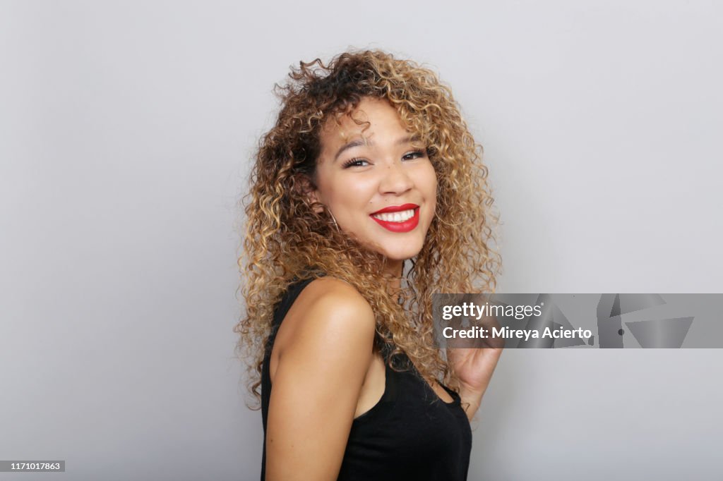 Portrait of young LatinX woman with curly blonde hair, wearing red lipstick and a black tank top, in front of a grey background.