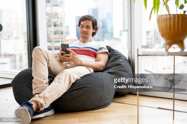 duizendjarige man ontspannen op een bean bag chatten met smartphone op een coworking office - beanbag chair stockfoto's en -beelden