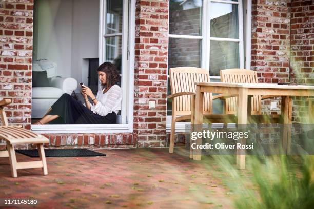 young woman sitting in windowframe using cell phone - french doors stockfoto's en -beelden