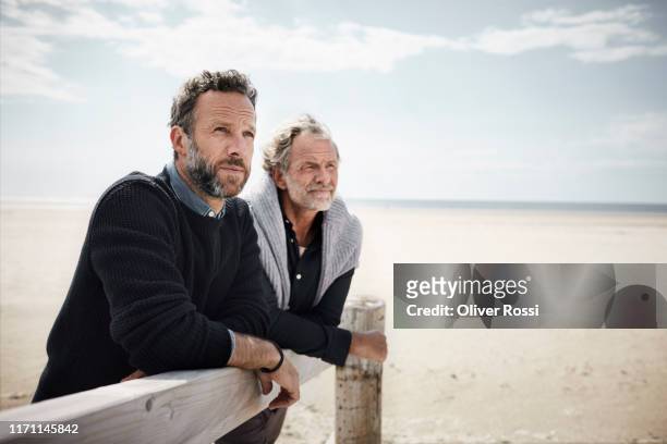 two mature men leaning on railing of boardwalk on the beach - familia de dos generaciones fotografías e imágenes de stock