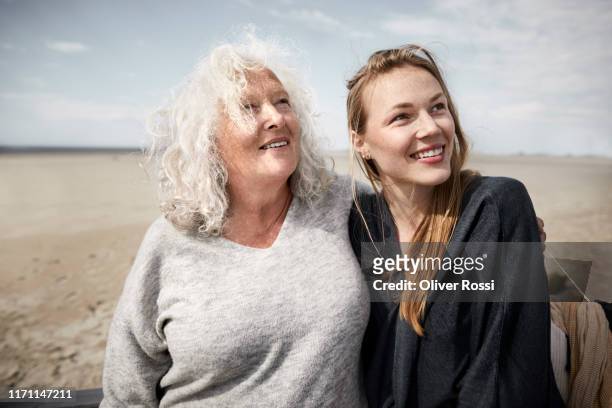 smiling mother with adult daughter on the beach looking sideways - mother and daughter in the wind stock-fotos und bilder