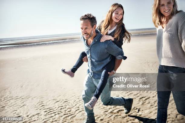 father carrying daughter piggyback on the beach - couple running on beach foto e immagini stock