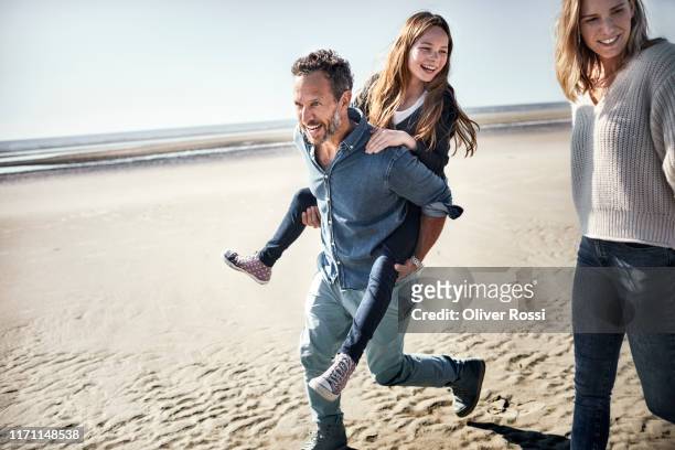 father carrying daughter piggyback on the beach - piggyback stock pictures, royalty-free photos & images