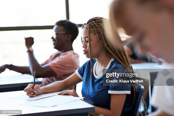 young woman writing exam at desk amidst friends - differential focus education stock pictures, royalty-free photos & images