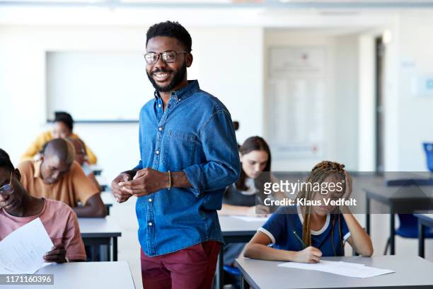 smiling supervisor amidst students writing exam - teacher stock-fotos und bilder