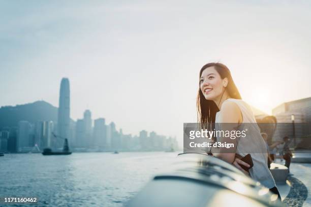 smiling young asian woman traveller looking out over promenade of victoria harbour enjoying the gentle breeze and the beautiful cityscape at dusk - hongkong lifestyle stockfoto's en -beelden