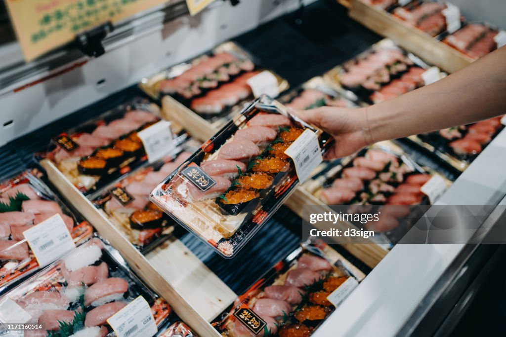 Woman's hand choosing a box of fresh packaged sushi from the display fridge in the supermarket