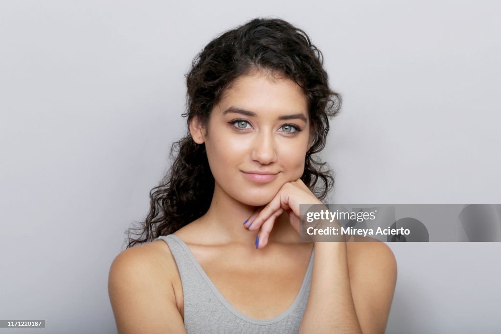 Portrait of young LatinX woman with curly brown hair and blue eyes, wearing a grey tank top in front of a grey background.