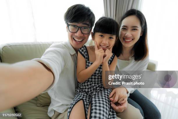 happy asian family with father, mother and daughter sitting and smile on sofa and taking selfie picture with smartphone at home - family smile stock-fotos und bilder