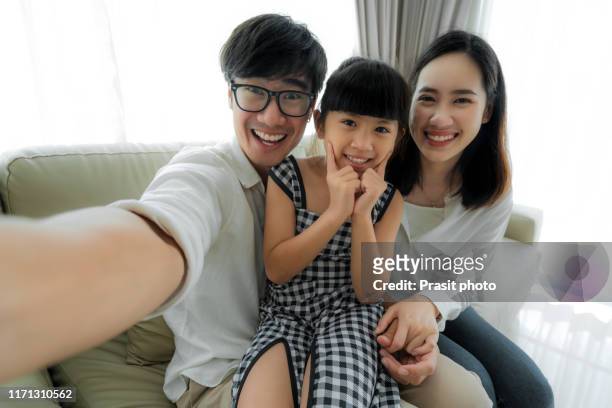 happy asian family with father, mother and daughter sitting and smile on sofa and taking selfie picture with smartphone at home - 自分撮り ストックフォトと画像
