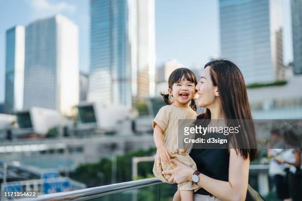 young asian mother and cute little daughter smiling joyfully over the urban terrace in downtown district against modern skyscrapers - prosperity stock pictures, royalty-free photos & images