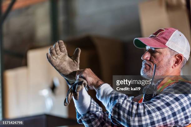 hispanic man standing in warehouse donning work gloves - trucker's hat stock pictures, royalty-free photos & images