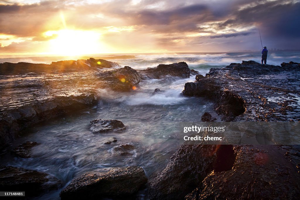 A fisherman on a rocky outcrop on the seashore at sunrise