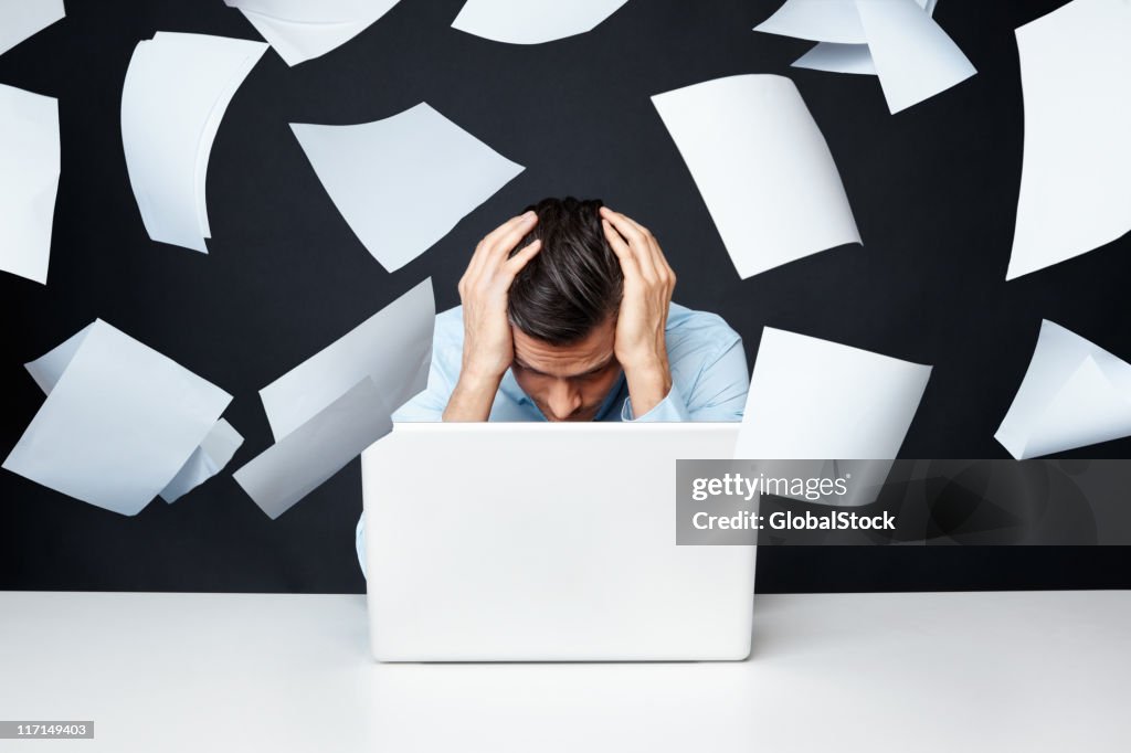 Man with hand on head looking at laptop with papers flying