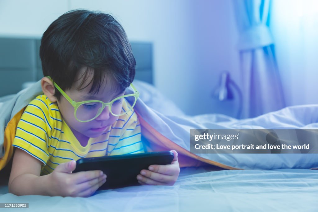 Schattig klein kind kijken film op smartphone op bed. Gevaren van blauw licht kunnen de ogen beschadigen. Knappe kleine jongen kan worden leeftijd gerelateerde macula degeneratie van blauw licht, draag brillen sinds de kindertijd