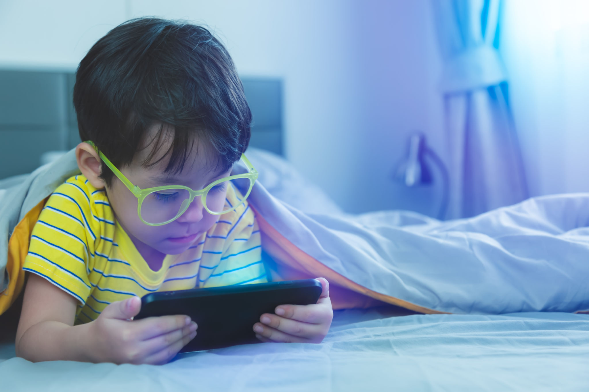 Cute little child watch movie on smartphone at bed. Dangers of blue light can damage eyes. Handsome little boy can be age related macular degeneration from blue light, wear eyeglasses since childhood Cute little child watch movie on smartphone at bed. Dangers of blue light can damage eyes. Handsome little boy can be age related macular degeneration from blue light, wear eyeglasses since childhood