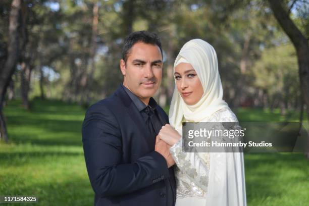 portrait of embraced muslim bride and groom couple looking at camera by holding each other's hands in a public park - arabic wedding stock pictures, royalty-free photos & images