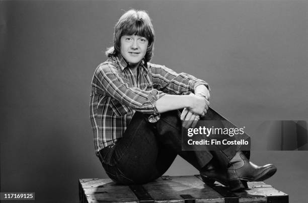 Keith Chegwin, Britiish television presenter, poses wearing a plaid shirt and sitting on a wooden topped table, in a studio portrait, 1978.
