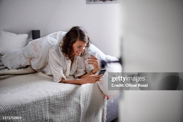 relaxed young woman drinking coffee and using cell phone in bed - nachtkleding stockfoto's en -beelden