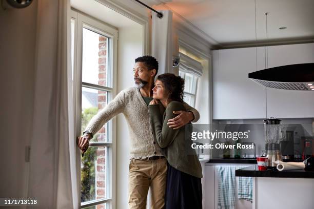 pensive man embracing young woman looking out of window in kitchen - smart windows stockfoto's en -beelden