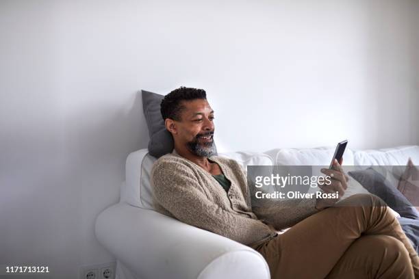 smiling man sitting on couch using cell phone - mann handy couch stock-fotos und bilder