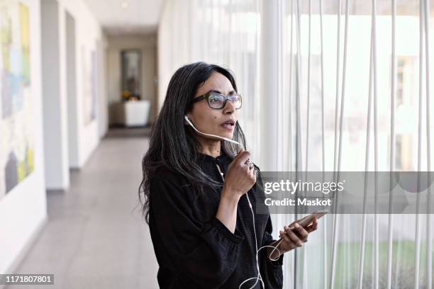 businesswoman talking on smart phone while standing by window in corridor at office - in ear headphones stock pictures, royalty-free photos & images