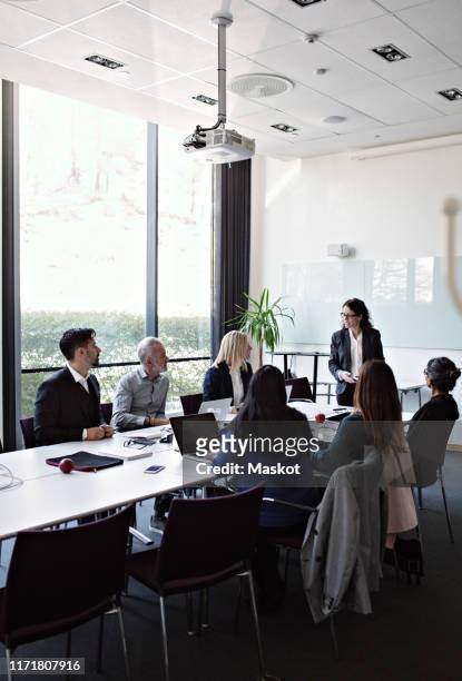 businesswoman interacting to female colleague sitting with coworkers at conference table during meeting in board room - formal businesswear stock pictures, royalty-free photos & images