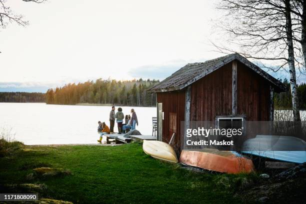 male and female friends talking outside cottage in front of lake during sunset - cottage stockfoto's en -beelden
